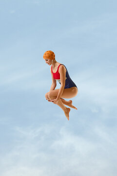 Orange capped athlete jumping with tucked knees against clouded sky backdrop with focused gaze. Sports motivation, movement training, active holidays concept.