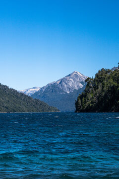 View of the deep blue waters of Nahuel Huapi Lake surrounded by lush forests and snow-capped mountains under a clear sky in San Carlos de Bariloche, Rio Negro, Argentina.
