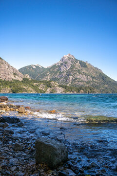 View of the rocky shoreline and turquoise waters of Nahuel Huapi Lake with the majestic Andes mountains in the background in San Carlos de Bariloche, Rio Negro, Argentina.
