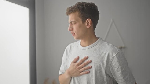 Young man holding hand to chest with closed eyes and pained expression in a building interior suggesting possible heartattack; discomfort concern.