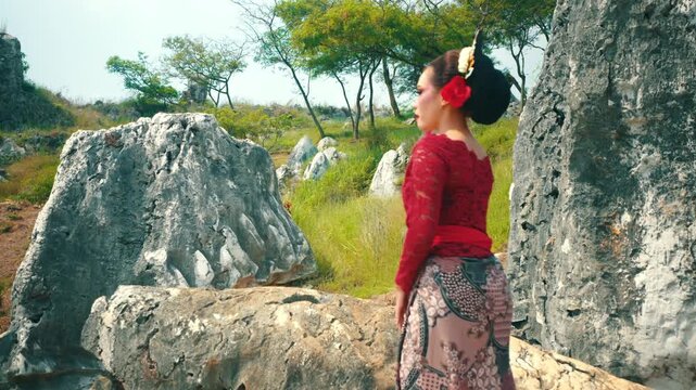 A medium shot capturing a woman in formal ethnic wear approaching a large rock to sit down and pose, surrounded by white stone formations and green trees.