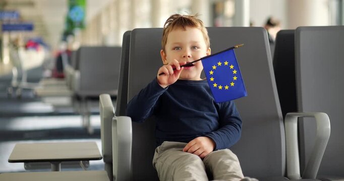 Little boy sits on airport terminal chair and waves small blue European Union flag. Smiling and moving hand with joyful expression, cheerful child speaks or sings during independent play while waiting