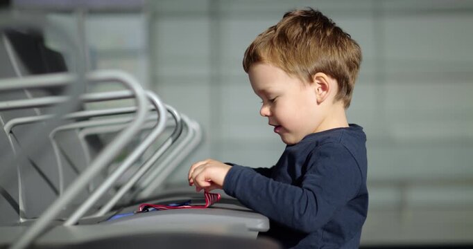 Little boy stands by airport terminal chairs and tries to fold tiny American flag. Concentrating deeply on fabric, child struggles playfully with small task during long flight delay waiting time.
