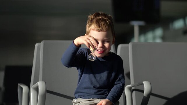 Little boy sits on airport terminal chair and curiously examines small glass hourglass. Smiling and laughing cheerfully as he inspects flowing sand, young child keeps himself busy during long wait