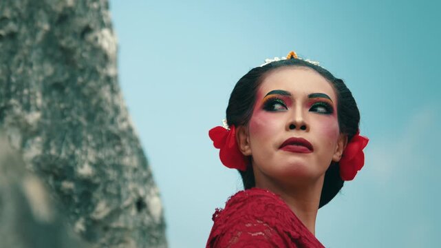 A low angle close-up portrait of a female dancer's face featuring bold traditional makeup, red lipstick, and hair accessories, looking confidently off-camera.
