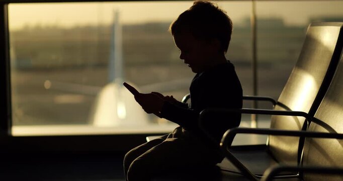 Silhouetted boy sits on against terminal window and interacts with smartphone. Tapping touchscreen with focus and curiosity, young child uses digital entertainment to pass time during flight delay.