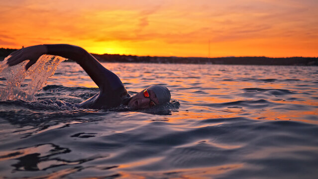 A swimmer male, training in open water during sunset. The determined athlete performs freestyle strokes, showcasing commitment to sport training and a healthy lifestyle.
