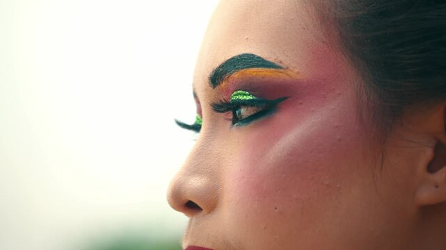 Macro profile shot capturing the detailed green glitter eyeshadow, thick eyebrows, and long false eyelashes of a female performer looking forward.