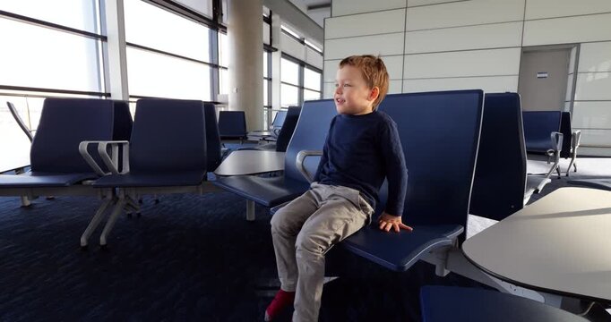 Sitting on lounge chair at airport terminal, active toddler dangles feet and raises hands while playfully speaking. Cheerful young child pauses to look aside during peaceful moment