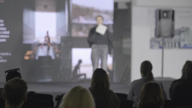 Seated attendees watch a speaker holding paper notes and a microphone before projected photographs.