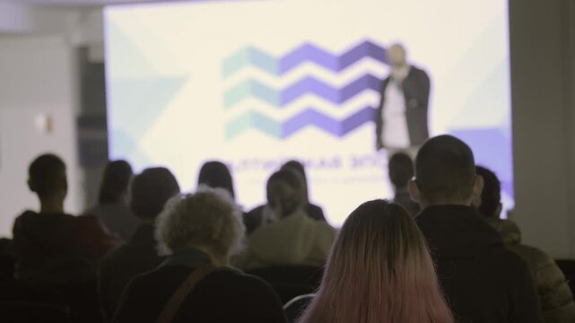 Audience silhouettes face a speaker before bright projected graphics, with rows of seated attendees in the foreground.