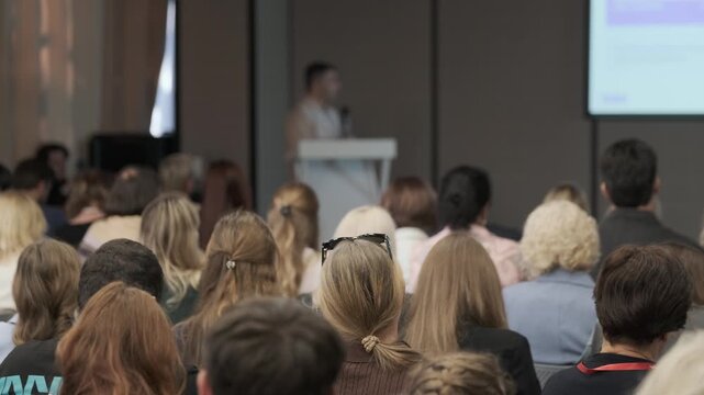 Conference seating fills the foreground during rightward camera panning toward projected slides, followed by mostly stationary framing.