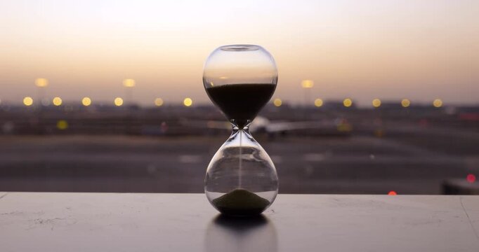 Trickling continuously inside transparent hourglass, dark sand measures passing time on airport terminal window sill, time-lapse shot. Blurry airplanes taxi, park, and land rapidly across airfield