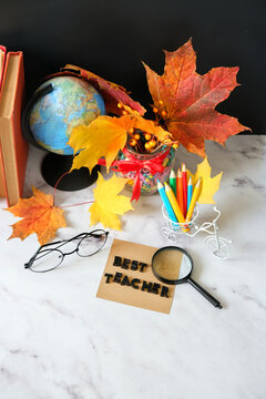 Teacher's day holiday concept. Paper card with text "Best teacher", school supplies, magnifying glass, autumnal leaves, eyeglass, globe and books on table close up. 
