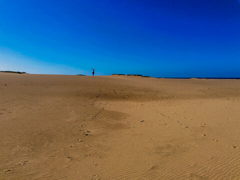 View of a person standing with arms raised on vast sand dunes under a clear blue sky in Saint Lucia, KwaZulu-Natal, South Africa.