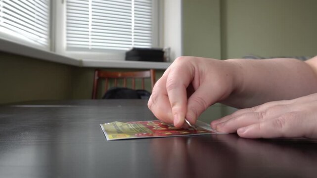 07.07.25. Russia. Moscow. A close-up of the hand of a man who uses a coin to erase the protective layer on a lottery ticket. The background shows a home interior with a window, blinds and furniture.
