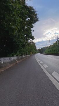 Vertical footage of a winding road through lush green forest, with chevron warning signs, concrete barriers, utility poles and a cloudy blue sky.