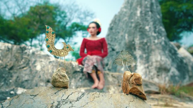 A creative composition showing traditional hair ornaments placed on rocks in the foreground, while an Indonesian woman in red Kebaya sits blurred in the background.
