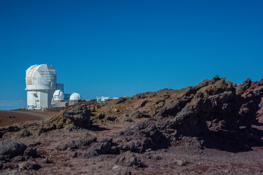 View of the Canada-France-Hawaii Telescope dome at the Mauna Kea Observatory surrounded by volcanic rocky terrain under a clear blue sky in Hawaii County, Hawaii, United States.