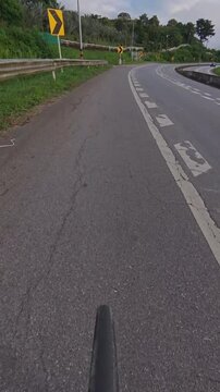 Vertical cyclist POV riding on a winding road through lush green forest, with chevron signs, guardrails and road markings along a scenic cycling route.