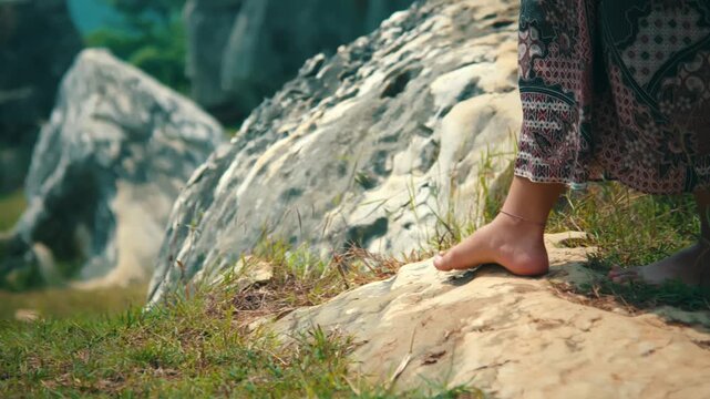 Close-up low angle tracking shot of a woman's bare feet walking carefully over grass and limestone rocks while wearing a traditional Batik skirt.