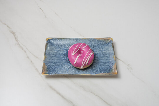 Pink strawberry-glazed mini donut with white icing drizzle on a rectangular blue ceramic plate over marble