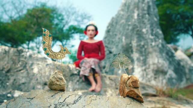 A wide shot focusing on a beautiful woman wearing a red Kebaya and Batik skirt sitting on a rock, framed by traditional hair accessories in the foreground.