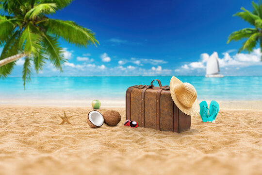 Vintage suitcase on sandy tropical beach with straw hat, flip flops, coconuts and starfish under palm trees, turquoise ocean and sailboat on horizon, summer vacation travel concept