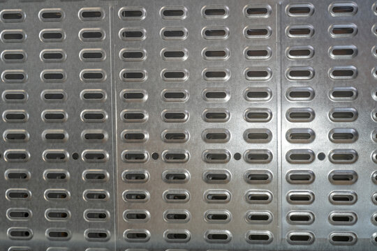 A gray metal surface features evenly spaced rows of elongated oval openings. Light and shadow emphasize the repeating industrial pattern and metallic texture.