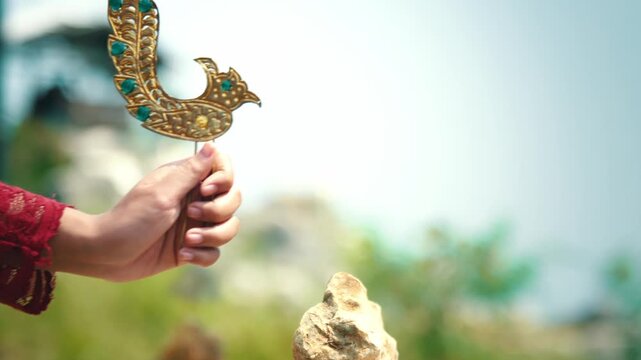 Close-up of a hand holding a golden peacock-shaped hair pin, a traditional Indonesian wedding accessory known as Cunduk Mentul.