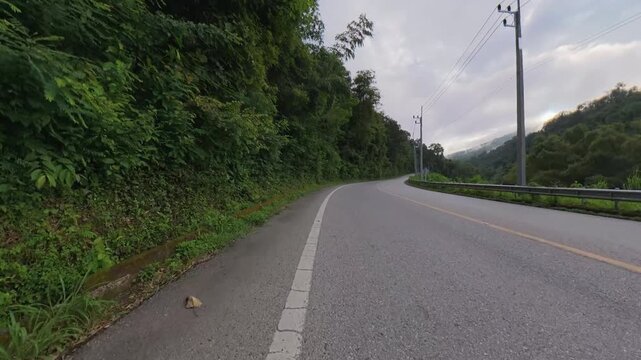 Scenic mountain road winding through lush green forest, with rock slopes, guardrails, utility poles and distant mountains under a cloudy sky.