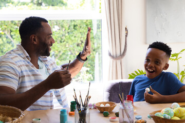 Obraz premium African American father and son painting eggs at wooden table by window, holding paintbrushes