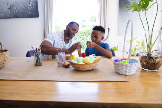 African American father and child painting colorful Easter eggs at dining table with paint jars