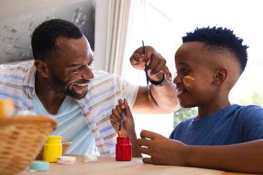 African American father in striped shirt and son painting at home table with paintbrushes and jars