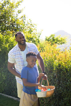 African American father and son smiling, holding wicker basket with bright eggs in sunny yard