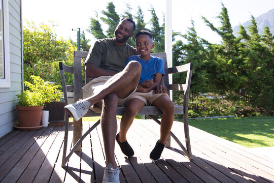 African American father and child sitting on wooden deck bench, smiling in shorts and sneakers