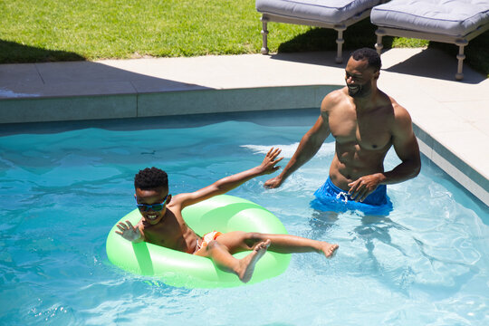African American father and son playing in backyard pool, child floating in lime-green ring