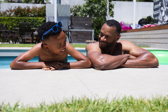 African American shirtless father-son leaning in backyard pool, smiling with blue shades