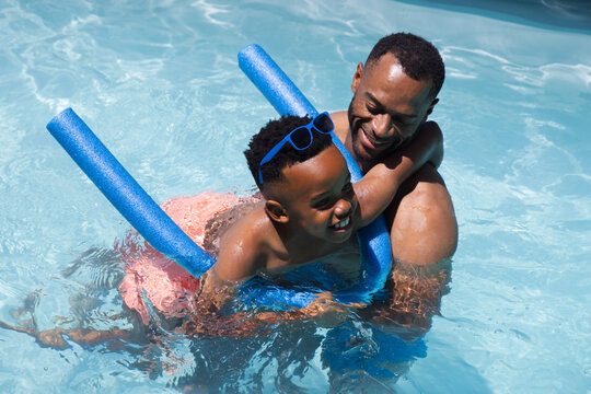 African American father and child wearing trunks, playing in pool with blue noodles and blue shades