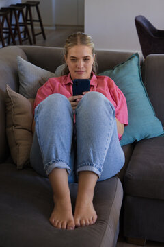 Woman sitting on gray sofa holding dark smartphone, wearing pink shirt and cuffed jeans
