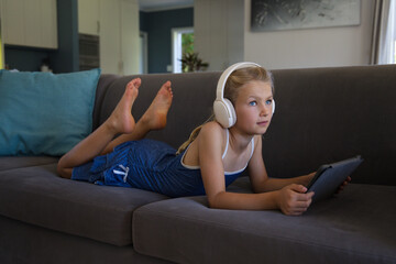 Female child lying on gray sectional sofa at home wearing blue dress and headphones holding tablet © WavebreakMediaMicro