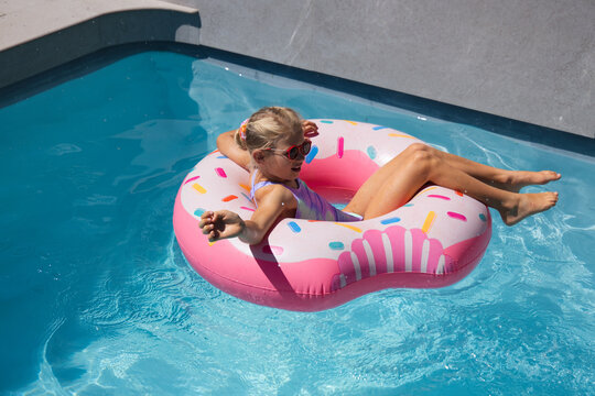 Child lounging on pink donut float in turquoise pool in swimsuit red shades holding dark object