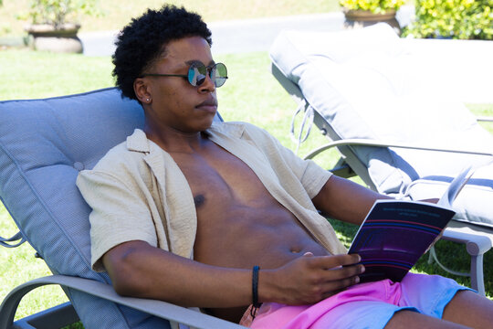 African American man reclining on lounger on green lawn wearing trunks and shades reading book