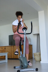 Obraz premium African American woman cycling on bike in living room pink tank holding phone dabbing with towel
