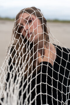 Woman lifting her chin beneath a fishing net at the beach. Mesh hangs across her face and black top as she angles her head against a blurred shore.