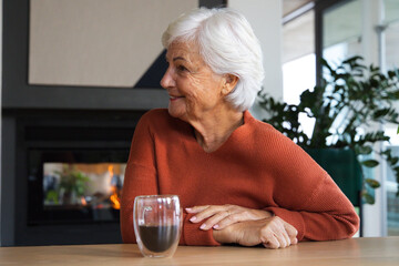 Obraz premium Senior woman sitting at table in lounge wearing rust sweater, holding glass mug, fireplace glowing