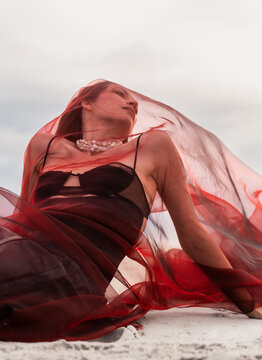 Woman beneath a red veil looking sideways from the sand. She reclines in a strappy top with pearl jewelry against an overcast sky.
