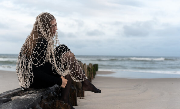 Woman seated on a beach groyne with net over her shoulders. She turns toward the camera with one knee raised against a backdrop of sea and sky.