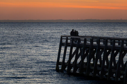 Luc-Sur-Mer, France - 08 08 2026:  Three black silhouettes of people wearing hoods are on a wooden pier in front of the sea, a colorful sunrise and a land far away