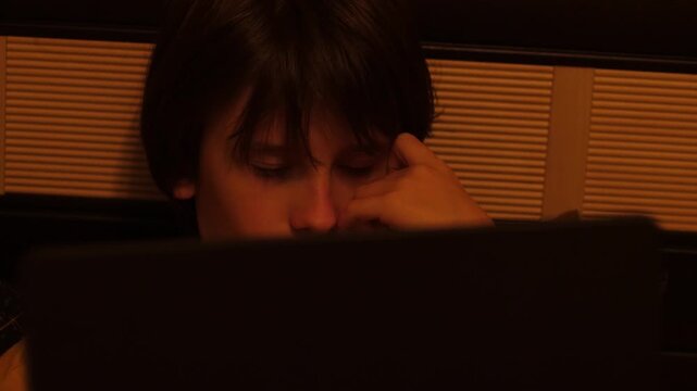 A teenage boy sits in front of a laptop screen in a dark room and attentively watches online content or studies. A young child spends the evening at home focused on the computer monitor